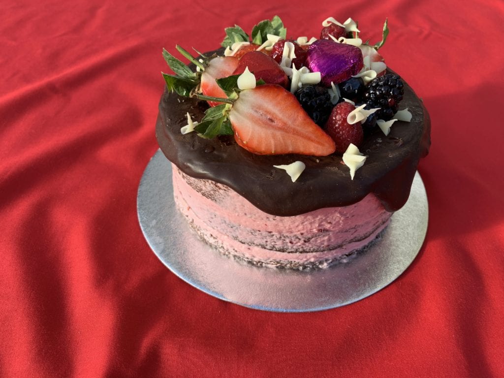 A round Valentines celebration cake with pink icing, chocolate top and fruit