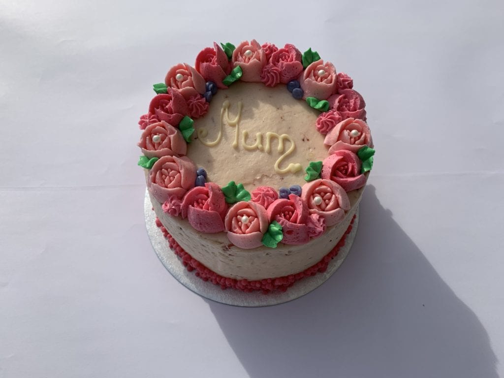 A round, iced and decorated mothers day cake with flower detailing and mum written in the centre made by quigleys bakery